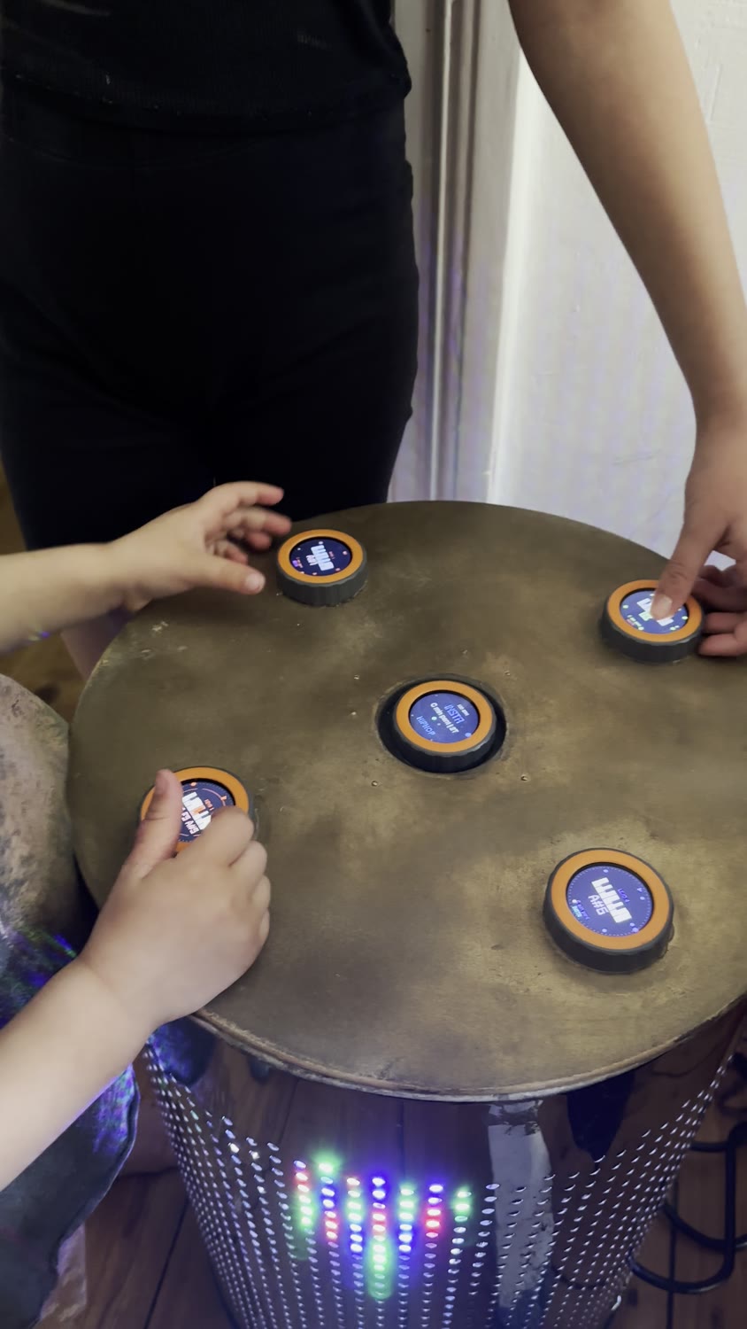 Zwei Personen spielen gemeinsam am runden TAMBUM-Tisch, im Sockel leuchten farbige Lichter